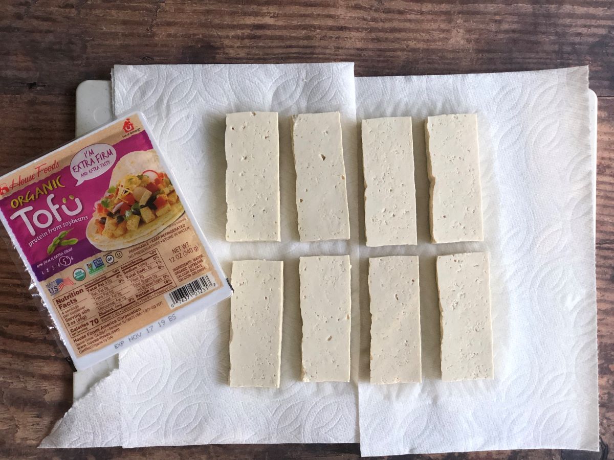 Block of tofu in it's packaging next to sliced tofu on a cutting board on a paper towel showing how to press the water out of tofu.