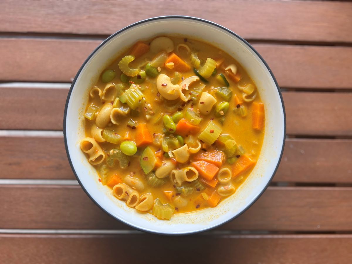 Bowl of coconut curry soup with celery, carrots, noodles, edamame, curry powder and coconut milk. Topped with red chili flakes.