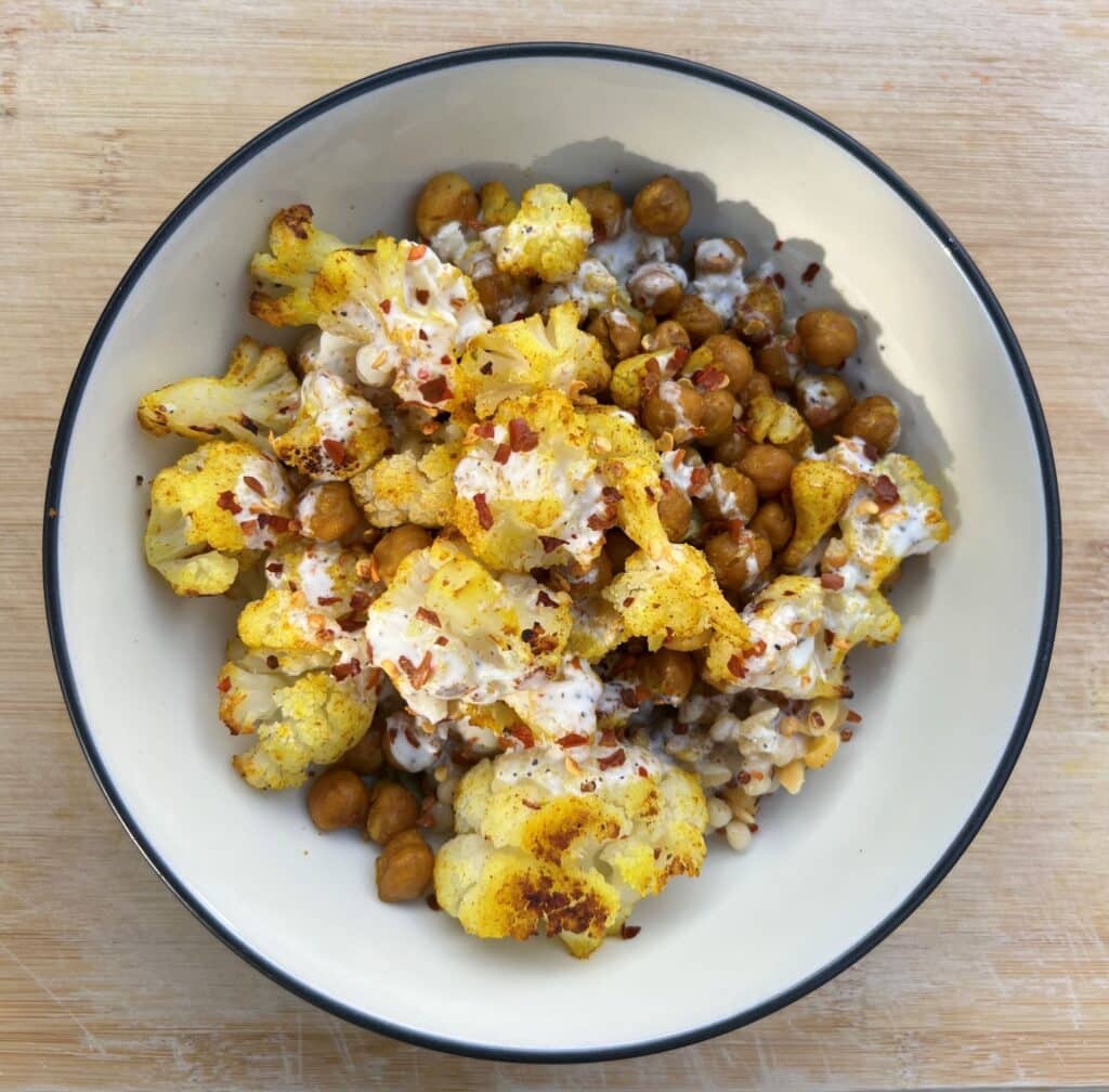Bowl of roasted chickpeas and cauliflower seasoned with curry powder and topped with yogurt dressing.