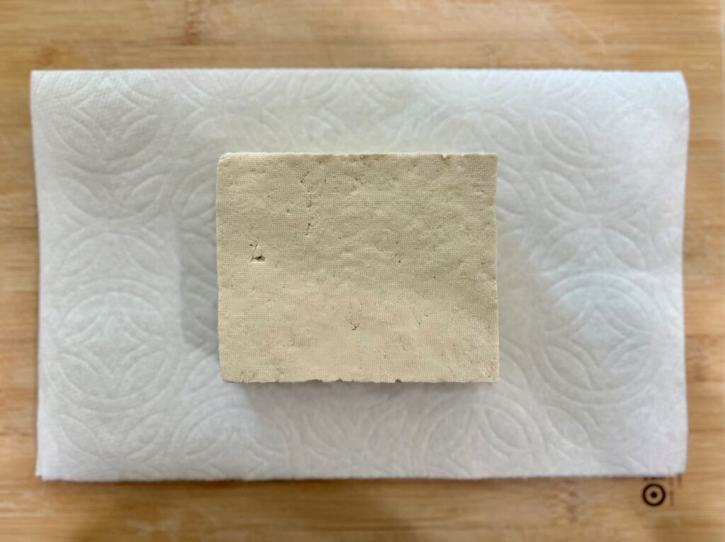 Block of tofu on a paper towel and cutting board getting ready to press the water out. 